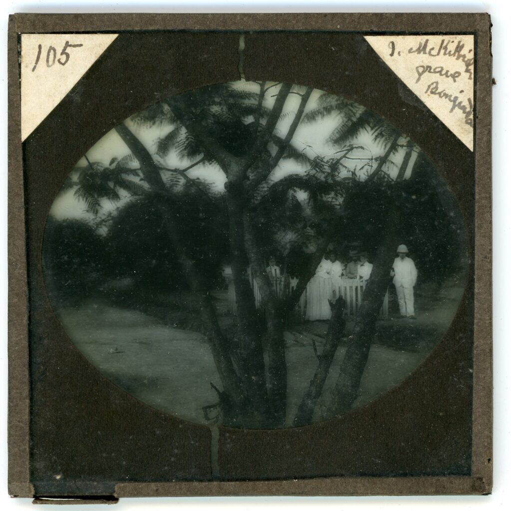 Missionaries gathered around a grave marked by a white picket fence