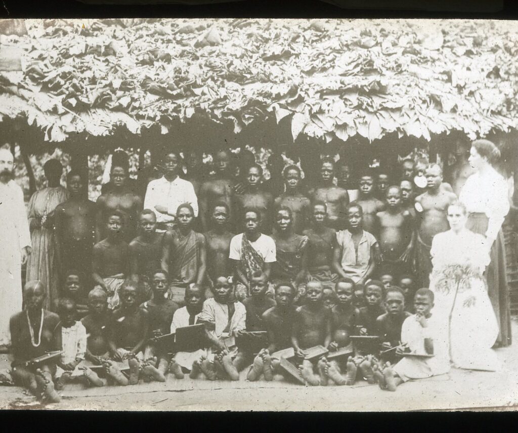 Local children and missionaries assembled under a school hut