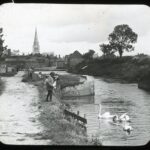 Slide showing the distant single spired tower of Chichester cathedral with a riverside path with Edwardian dressed children feeding swans
