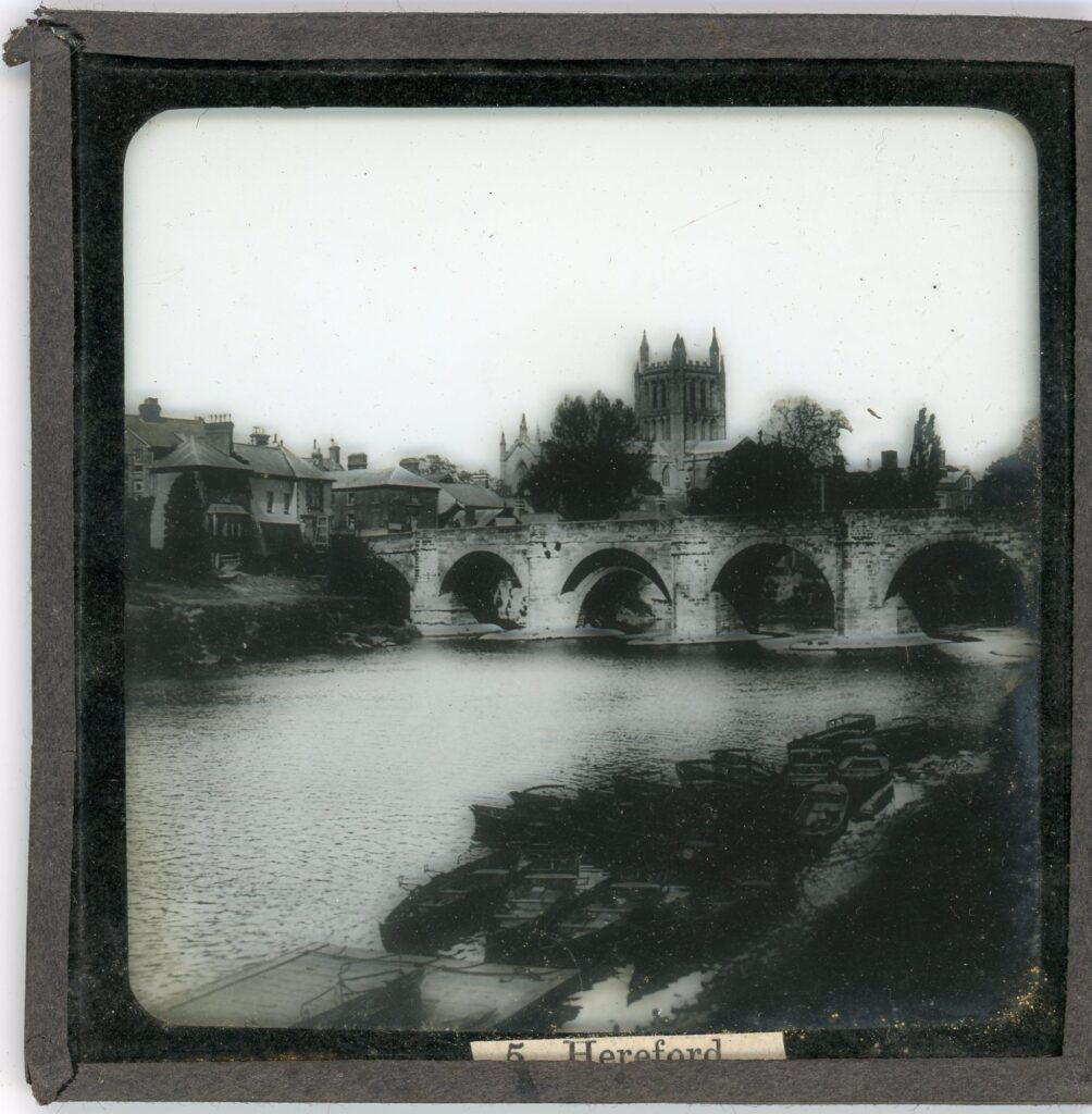 Side showing the bell tower of Hereford cathedral as seen from across a river with numeroud rowing boats and crossed over by a stone, 5 piered medieval bridge