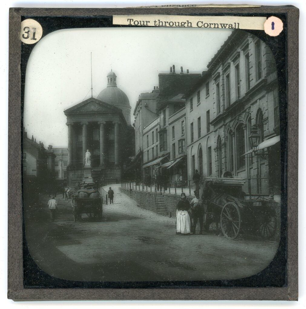 Slide showing a wide road of terraced commercial buildings with raised pedestrian path and neo-classical hall, along with numerous horse-drawn carriages and Edwardian attired people.