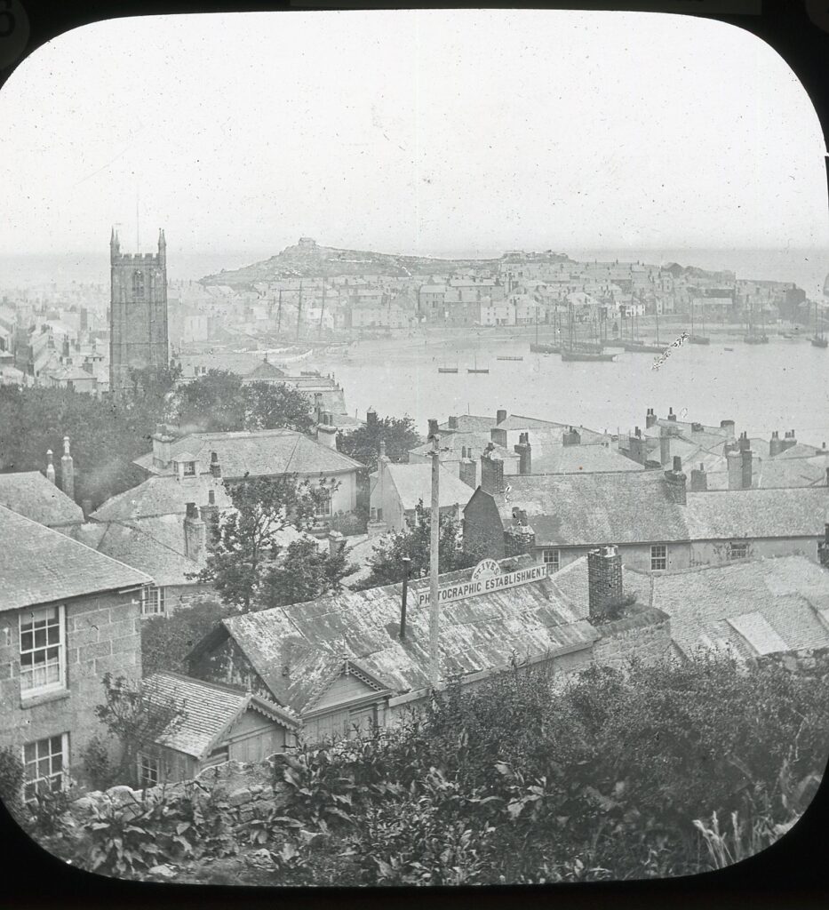 Slide showing a view over tiled rooftops and chimneys to a curved beach and harbour with moored ships and prominent church tower