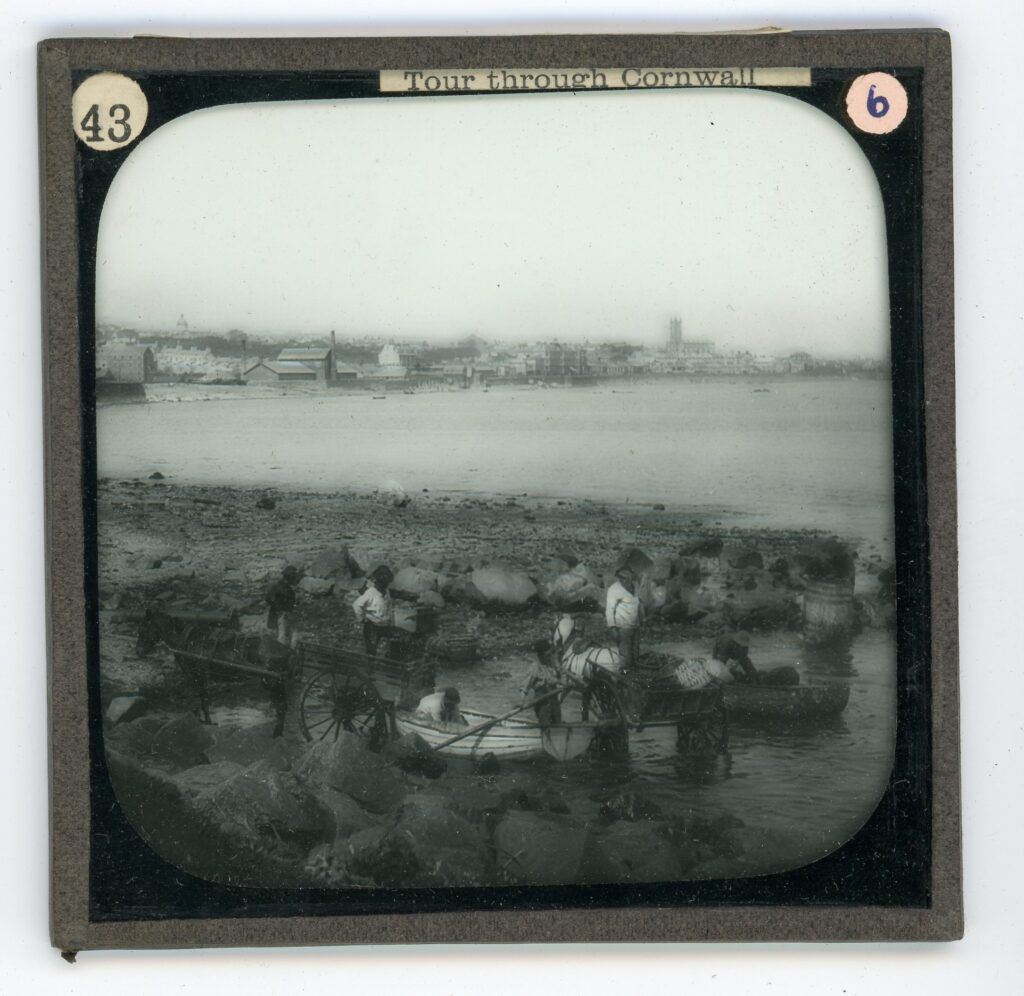 Slide showing row boats being unloaded into horse drawn carts in shallow water with a stretching view of urban coastline behind