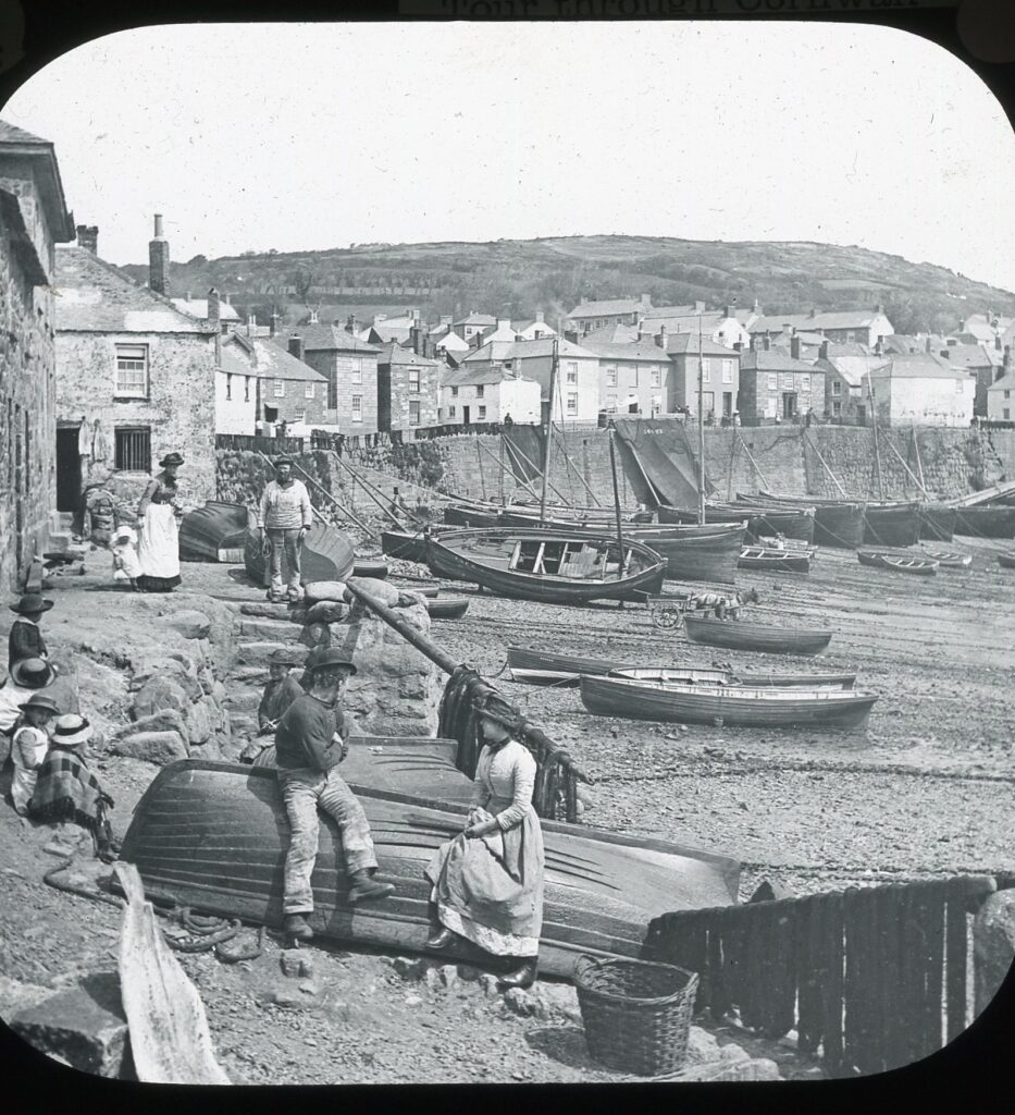 Slide showing Edwardian clad men, women, and children resting on a harbour wall at low tide with a view of waterside cottages and moored boats resting on the shore