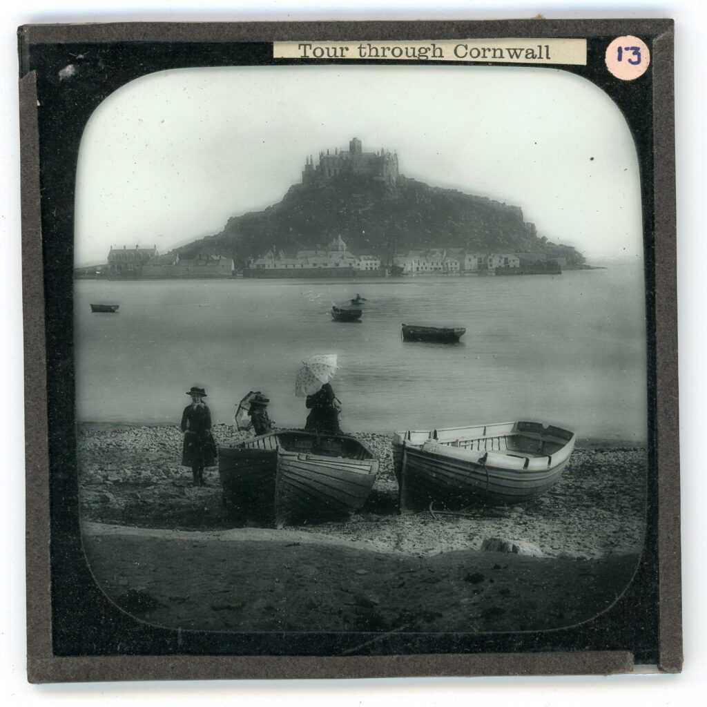 Slide showing a view of a near island with a gothic church atop, with row boats and Edwardian clad woman and children with parasols in the foreground