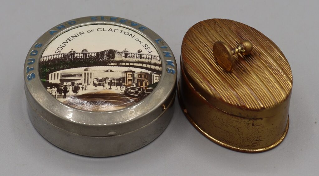 two boxes, one round silver metal with a picture on the lid, the other is oval gold coloured metal with a collar stud on the lid