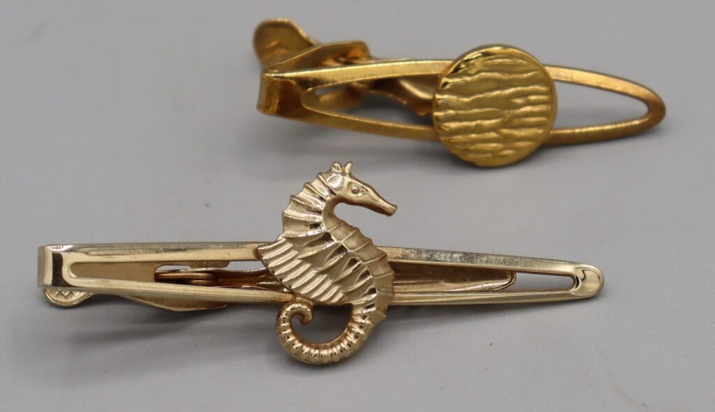 Two tie clips, gold tone metal. One has a seahorse on the centre, the other has a round textured disc.